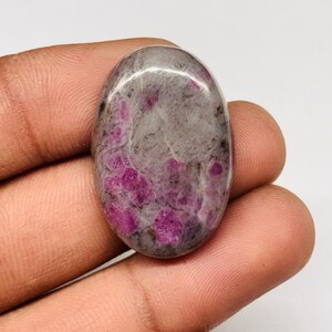 Peut inclure: Une pierre précieuse de forme ovale avec une surface polie. La pierre présente un mélange de teintes grises et blanches, avec des inclusions magenta vives dispersées. La pierre est tenue dans une main, mettant en valeur sa taille et sa texture.