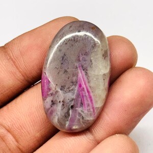 Peut inclure: Pierre précieuse ovale à surface polie. La pierre présente un mélange de couleurs, notamment du gris, du blanc et des stries roses vives. La pierre est tenue dans une main, mettant en valeur sa taille et sa forme.