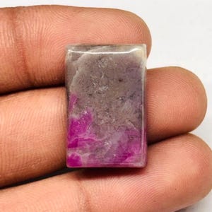 Peut inclure: Une pierre précieuse rectangulaire avec un mélange de couleurs. La partie supérieure est gris clair, passant à un gris plus foncé, puis à un magenta vif en bas. La pierre a une surface polie.