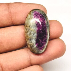 Peut inclure: Une pierre précieuse de forme ovale avec un contraste de couleurs saisissant. La pierre présente une teinte magenta profond et vibrante d'un côté, passant à un mélange de gris, de brun et de blanc de l'autre. La surface polie met en valeur les motifs naturels.