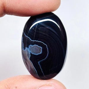 以下が含まれることがあります： 楕円形の研磨された宝石。濃いネイビーブルーのベースに、渦巻く白と水色の模様が施されています。滑らかで反射する表面を見せるため、石は指で持たれています。デザインは目に似ています。