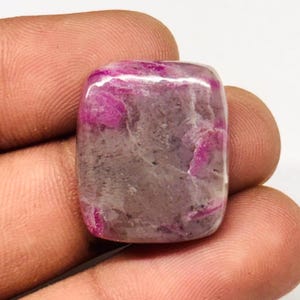 Peut inclure: Une pierre précieuse rectangulaire polie avec un mélange de teintes roses et grises. La pierre a une surface lisse et arrondie et semble translucide, avec des motifs internes visibles. La pierre est tenue dans une main.