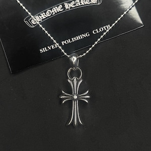 May include: A silver pendant necklace featuring a cross design with a small gemstone at the center. The necklace is displayed on a black surface with a card that reads "CHROME HEARTS SILVER POLISHING CLOTH".