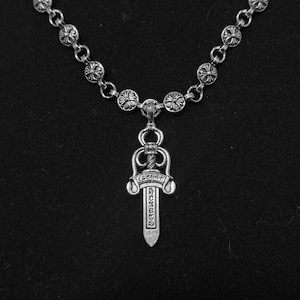 Puede incluir: Collar plateado con un colgante en forma de daga. El colgante tiene detalles ornamentados y texto. La cadena está compuesta por pequeños eslabones circulares decorativos. El collar se muestra sobre un fondo negro.