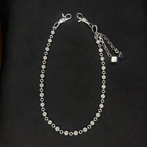 May include: A silver-colored wallet chain with a floral design. The chain features small, round, clear stones set in a floral pattern. The chain has two clasps on either end and a small dangling charm with a cube.