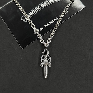 Puede incluir: Un collar plateado con un colgante de daga. El colgante tiene detalles intrincados y está suspendido de una cadena. El collar se muestra sobre una superficie negra con una tarjeta que dice "Chrome Hearts".