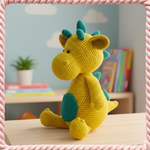 May include: A handmade yellow crochet dragon with teal accents. The plush toy has a round head, small black eyes, and teal spikes along its back and head. A teal circle is on the dragon's side. The dragon is sitting on a light brown surface.
