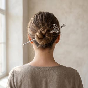 May include: A silver-toned metal hair pin shaped like a sword with an ornate, filigree-style hilt and a pointed blade. The hair accessory is inserted horizontally through a neat, round bun of light brown hair, securing the style in place.