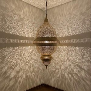 May include: An elaborate, gold-toned pendant lamp with detailed cut-out designs. The lamp is suspended from a chain and projects a decorative light pattern onto the walls. The lamp has a rounded form with a pointed base.