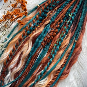 May include: A collection of colorful dreadlocks in shades of teal, copper, brown, and blonde. Some dreads are braided, others are twisted, and some are adorned with beads. The dreadlocks are arranged on a white, fluffy surface, with dried orange flowers in the upper left corner.