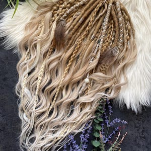 Könnte beinhalten: Ein Set aus blonden und hellbraunen Dreadlocks mit welligen Enden. Die Dreadlocks sind mit silbernen Blattanhängern, Federn und Perlenakzenten verziert. Das Haar ist mit Zöpfen und losen Strähnen gestylt.