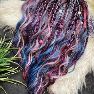 Puede incluir: Una colección de rastas coloridas en tonos azul, rosa, morado y gris. Las rastas están adornadas con cuentas, abalorios y cintas. El cabello está peinado sobre una superficie blanca y esponjosa.