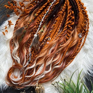 May include: Handmade dreadlocks in shades of brown, orange, and blonde, decorated with feathers, beads, and white lace. The dreadlocks are arranged on a white, fluffy surface, with dried flowers and greenery in the background.