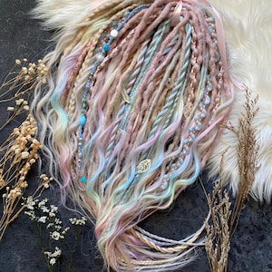 Puede incluir: Un conjunto de rastas de colores pastel con tonos rosas, azules y amarillos. Las rastas están adornadas con cuentas, abalorios y detalles de hojas metálicas. Una cinta rosa está atada en la parte superior. Las rastas están peinadas sobre una superficie blanca similar a la piel.
