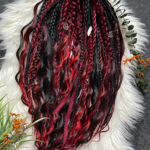 Peut inclure: Ensemble de dreadlocks noirs et bordeaux avec des touches de rouge et des ornements de feuilles argentées. Les dreadlocks sont coiffés avec un mélange de tresses et de mèches lâches, attachés avec un ruban rouge. Présenté sur une surface en fausse fourrure blanche.