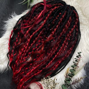 Op de afbeelding: Zwarte en rode dreadlocks met een gradiënteffect, versierd met rode linten en kleine kralen. Een hartvormige bedel is bevestigd. Het haar is gestyled op een witte, zachte ondergrond. Een opvallende look.
