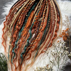 Puede incluir: Conjunto de rastas hechas a mano en tonos marrones, naranjas y turquesas. Las rastas están peinadas con trenzas y torzadas, adornadas con pequeñas cuentas y un dije de hoja decorativo. Dispuestas sobre una superficie blanca y esponjosa.