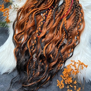 Puede incluir: Una colección de rastas naranjas, marrones y negras, peinadas con trenzas y secciones onduladas. Las rastas están adornadas con pequeñas cuentas y abalorios decorativos, y atadas con una cinta roja. El cabello está dispuesto sobre una superficie blanca y esponjosa.