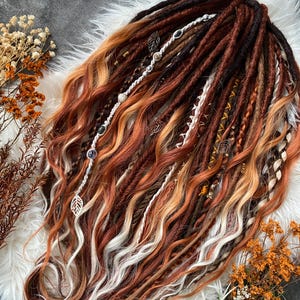 Può includere: Una collezione di dreadlocks fatti a mano nei toni del marrone, arancione e biondo. I dread sono adornati con perline decorative e ciondoli a forma di foglia. I capelli sono acconciati su una superficie bianca e soffice.