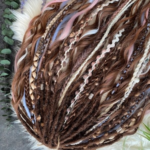 May include: A collection of brown, white, pink, and light blue dreadlocks. The dreadlocks are styled with braids and decorative beads. The hair is arranged on a white fluffy surface with greenery and purple flowers in the background.