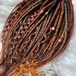 May include: A collection of handmade dreadlocks in shades of brown, copper, and black. The dreads are adorned with decorative beads and metal accents. Some dreads are braided, and others are wrapped with thread. The overall style is bohemian and natural.