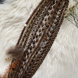 May include: Brown and tan dreadlocks with decorative accents. The dreadlocks are styled with white and tan braiding, wooden beads, silver leaf charms, and a feather. The hair accessory is arranged on a white, fluffy surface.