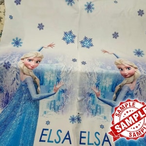 Op de afbeelding: Witte stof met een blauwogig stripfiguur in een blauwe jurk, met het woord "ELSA" in blauw. Het ontwerp bevat blauwe sneeuwvlokken en een waterval achtergrond. Een rode "SAMPLE" stempel is zichtbaar.