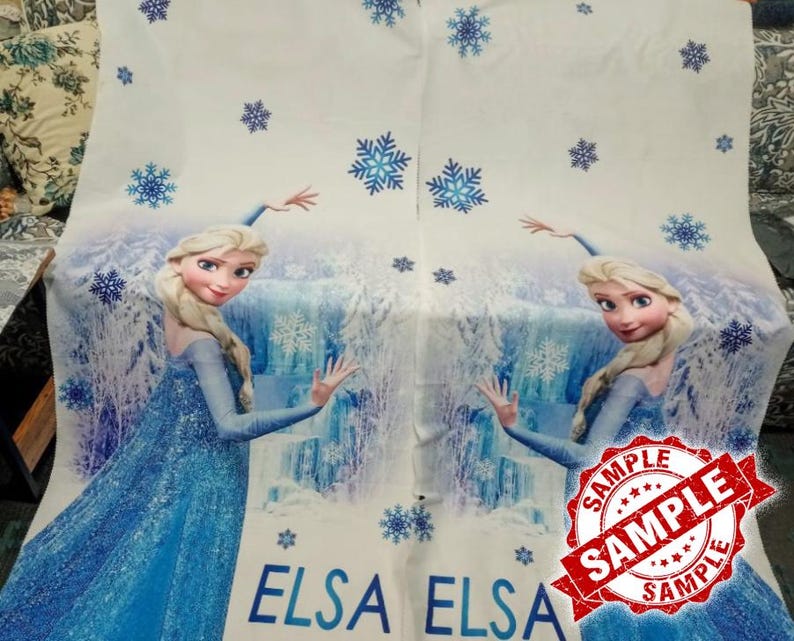 Op de afbeelding: Witte stof met een blauwogig stripfiguur in een blauwe jurk, met het woord "ELSA" in blauw. Het ontwerp bevat blauwe sneeuwvlokken en een waterval achtergrond. Een rode "SAMPLE" stempel is zichtbaar.