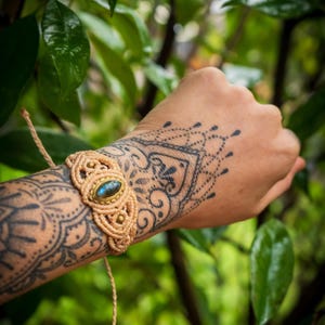 Könnte beinhalten: Ein beigefarbenes Makramee-Armband mit einem ovalen Stein, wahrscheinlich Labradorit, in einem goldfarbenen Rahmen. Das Armband wird an einem Arm mit aufwendigen Tätowierungen getragen. Der Hintergrund ist ein verschwommenes grünes Laub.