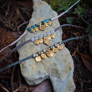 Könnte beinhalten: Drei handgefertigte Armbänder mit goldfarbenen Charms und Perlen liegen auf einem hellen Stein. Die Armbänder haben unterschiedlich farbige Kordeln: hellrosa, grau und türkis. Die Charms umfassen Münzen, Quadrate und ein Handsymbol.