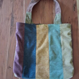 Op de afbeelding: Een veelkleurige tote bag met een suède-achtige textuur. De tas heeft verticale strepen in tinten paars, marineblauw, goud, blauwgroen en olijfgroen. De handvatten zijn een gedempte bruine kleur. De tas staat op een houten oppervlak.