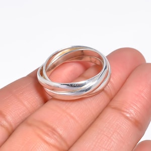 May include: A silver ring with three interlocking bands. The bands are smooth and polished, reflecting light. The ring is shown on a hand against a white background. A piece of jewelry.