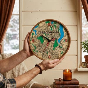 May include: A round wooden wall clock featuring a map of Sevastopol. The clock face has a beige background with green, blue, and red accents. The clock hands are black, and the numbers are red. The clock is held by a person.