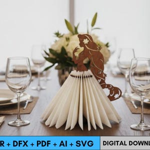 May include: A brown laser-cut mermaid-shaped napkin holder with a fan of folded white napkins. The mermaid is positioned in front of a bouquet of white flowers on a wooden table. Wine glasses and plates are set for a formal dinner.