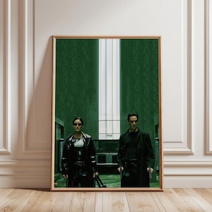 May include: Framed poster featuring a scene from the movie "The Matrix." The image shows two figures in black outfits, standing in a green-toned room. The poster is in a wooden frame and leans against a white wall.