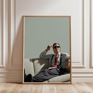 Könnte beinhalten: Gerahmter Druck mit einem Mann mit Sonnenbrille, der auf einem grauen Sofa sitzt. Er trägt ein gestreiftes Hemd, eine rote Krawatte, Hosenträger und eine Nadelstreifenhose. Das Kunstwerk wird vor einem hellgrauen Hintergrund gezeigt.