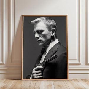 May include: Black and white framed print of a man in a tuxedo holding a handgun. The man is looking to the side with a serious expression. The print is in a wooden frame and leans against a white wall.