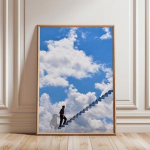 May include: A framed art print depicting a person climbing a staircase that merges into a bright blue sky filled with white clouds. The person is dressed in black. The artwork is displayed on a wooden floor.
