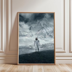 Puede incluir: Impresión enmarcada que presenta a un astronauta con un traje espacial blanco, de pie sobre una superficie oscura con montañas nevadas y un cielo nublado. La obra de arte se muestra en un marco de madera.