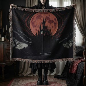 May include: Black woven throw blanket featuring a gothic castle silhouette under a large red moon. The blanket has a fringed edge and a dark, dramatic aesthetic. The design evokes a spooky, fantasy theme.