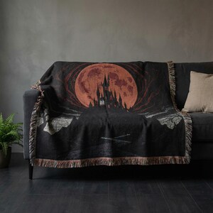 May include: Black throw blanket featuring a gothic castle silhouette against a large, reddish-orange full moon. The blanket has a fringed edge and is draped over a dark gray sofa. A beige pillow is on the sofa.
