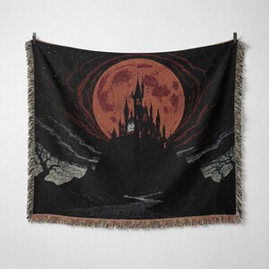 May include: Black throw blanket featuring a gothic castle silhouette against a large, reddish-orange full moon. The design includes a dark night sky with stars and a river. The blanket has a fringed edge.