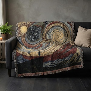 May include: A woven throw blanket featuring a cosmic design with a swirling galaxy, a sun, and a figure standing on a cliff. The color palette includes gold, red, blue, and black, with a fringed edge. The blanket is draped over a dark gray sofa.