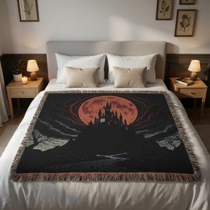 May include: A black throw blanket featuring a spooky castle silhouette against a large orange moon. The design includes dark clouds and a detailed castle, with a fringed edge. The blanket is displayed on a bed with neutral-toned pillows.