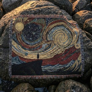 May include: A woven throw blanket featuring a stylized depiction of a starry night with a figure standing on a cliff. The design incorporates swirling patterns in shades of gold, blue, red, and black, with a fringed edge.