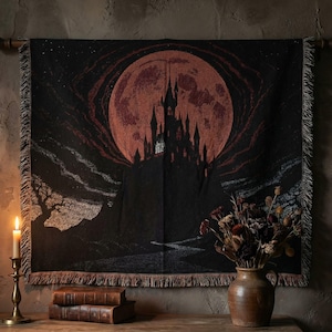 May include: Black tapestry featuring a gothic castle silhouette against a large, reddish-orange moon. The tapestry has a fringed edge and is hanging on a wooden rod. A lit candle and a vase of dried flowers are in the foreground.