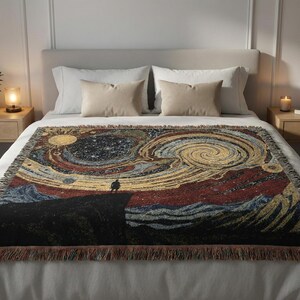 May include: A woven throw blanket with a colorful, abstract design depicting a starry night sky and a figure on a cliff. The blanket features a fringed edge and is displayed on a bed with pillows.