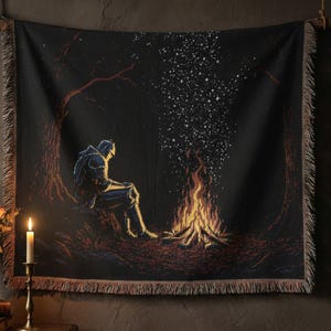 May include: Black woven throw blanket featuring a knight sitting by a campfire under a starry night sky. The artwork has a pixelated style with a fringed edge. The color palette includes black, brown, orange, and white.