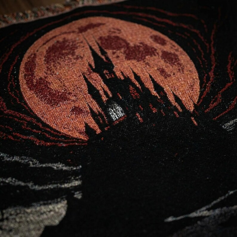 May include: A woven tapestry featuring a gothic castle silhouette against a large, reddish-orange full moon. The castle is black, with detailed spires and towers. The moon has a textured appearance, and the background is dark.