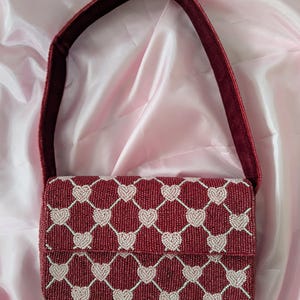 May include: A burgundy beaded handbag featuring a pattern of white hearts. The rectangular bag has a flap closure and a matching burgundy strap. The background is a pink satin fabric.